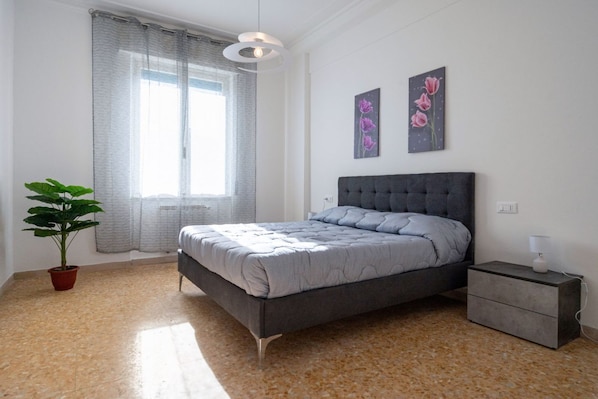 Un dormitorio con cama, mesita de noche, planta en maceta y ventana con cortinas.