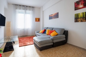 Una sala de estar con un sofá gris, una mesa de centro de vidrio, un tapete rojo y obras de arte en la pared.