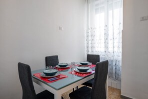 Un comedor con una mesa dispuesta para cuatro personas, sillas, una ventana con cortinas translúcidas y un interruptor de luz en la pared.