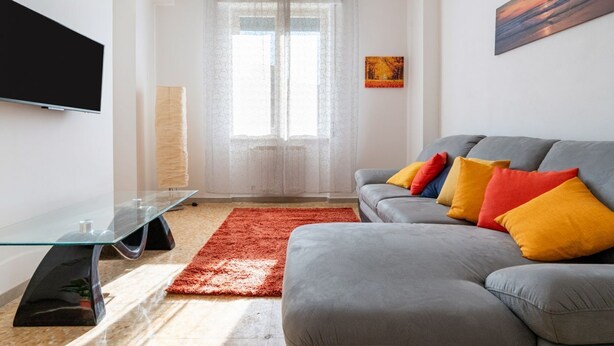 Un salon moderne avec un canapé gris, des coussins colorés, une table basse en verre, un téléviseur à écran plat et une grande fenêtre ornée de rideaux légers.