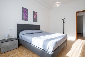 A bedroom with a grey upholstered bed, a bedside table with a lamp, and two wall-mounted flower paintings. - GuestHost - Bright, newly renovated apartment, which comfortably accommodates up to 6 people. Perfectly positioned close to essential services and the city centre, the accommodation is close to the city's main square and a few minutes from the ferry term (La Spezia)