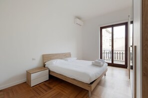A bedroom with a wooden bed, a nightstand, a white drawer, a window with a view of buildings, and a sliding door to an outdoor balcony.