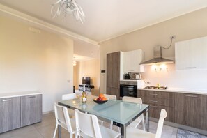 A modern kitchen with a glass dining table, white chairs, and a bowl of fruit.