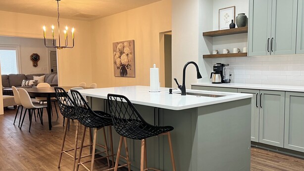 A modern kitchen with a central island, bar stools, a chandelier, and a dining area with a sofa and chairs.