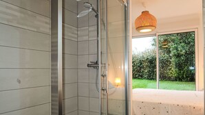 Een moderne badkamer met een glazen douchecabine, een bed met een witte spreidekker en een groot raam met uitzicht op groen buiten.