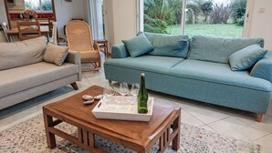 Een woonkamer met een bank, een houten bijzettafeltje met een fles en glazen, een rieten fauteuil en uitzicht op een tuin door grote ramen.