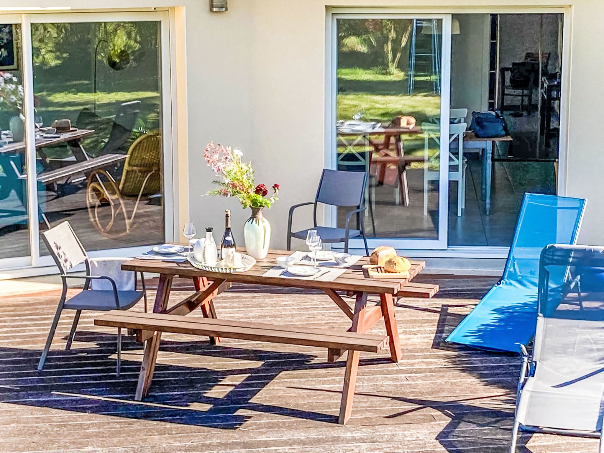 An outdoor dining area with a table set for two, a vase with flowers, and a view of the garden through glass doors.