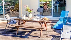 Een buitenterras met een gedekte tafel voor twee, een vaas met bloemen en uitzicht op de tuin door glazen deuren.