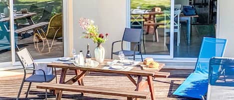 An outdoor dining area with a table set for two, a vase with flowers, and a view of the garden through glass doors.