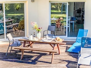 An outdoor dining area with a table set for two, a vase with flowers, and a view of the garden through glass doors.