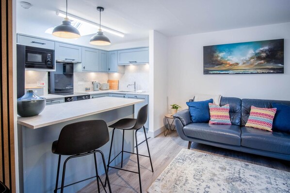 A modern kitchen with a central island, bar stools, and a dining area with a sofa and cushions. - Sandy Steps - 1 Bedroom Apartment - Tenby (Tenby)