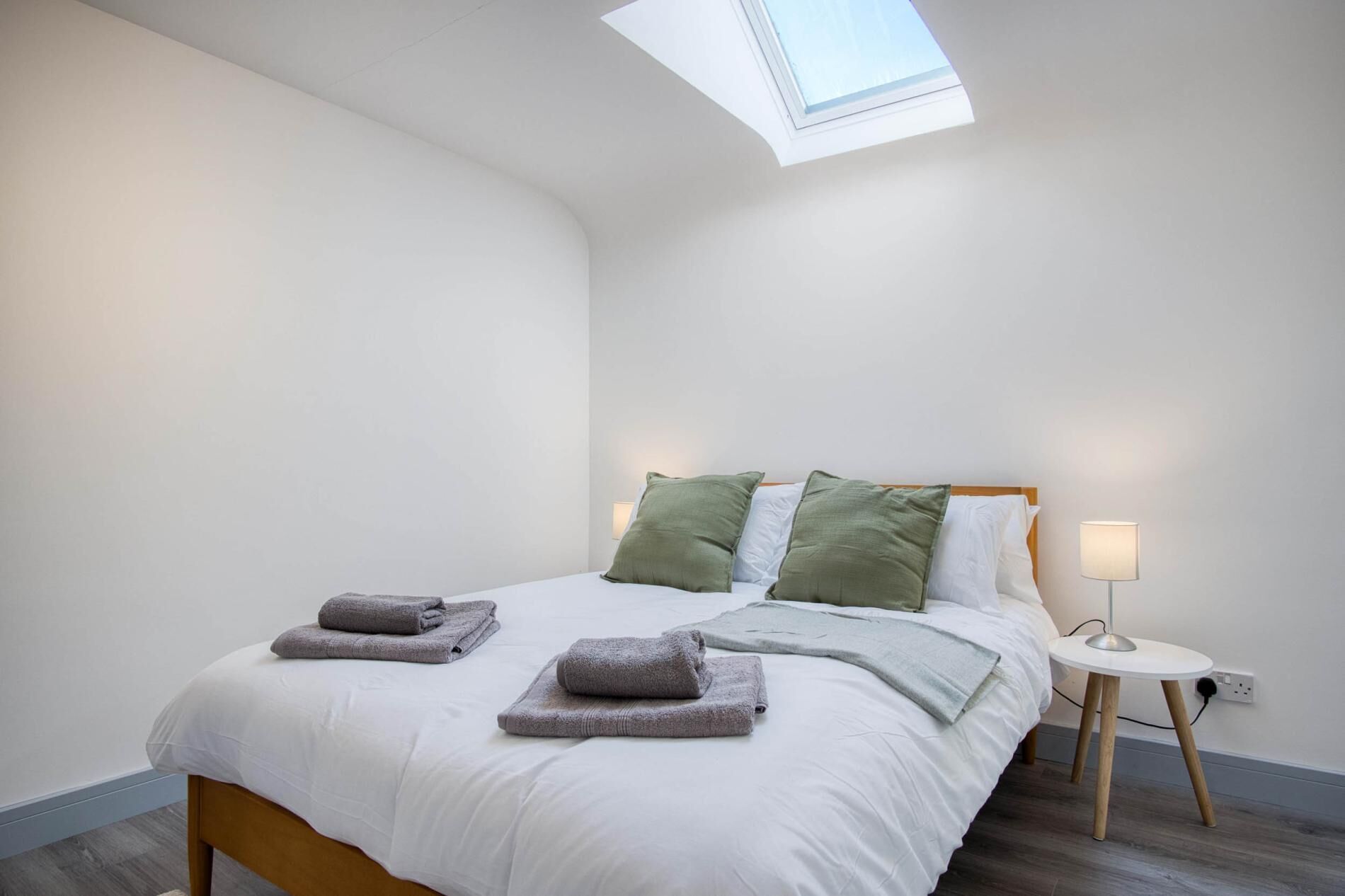 Un lit bien fait, avec du linge de lit blanc et deux serviettes pliées, une lampe de chevet et une lucarne au-dessus du lit.