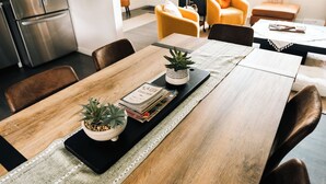 A dining area with a wooden table, chairs, a black tray with books and plants, and a kitchen in the background.