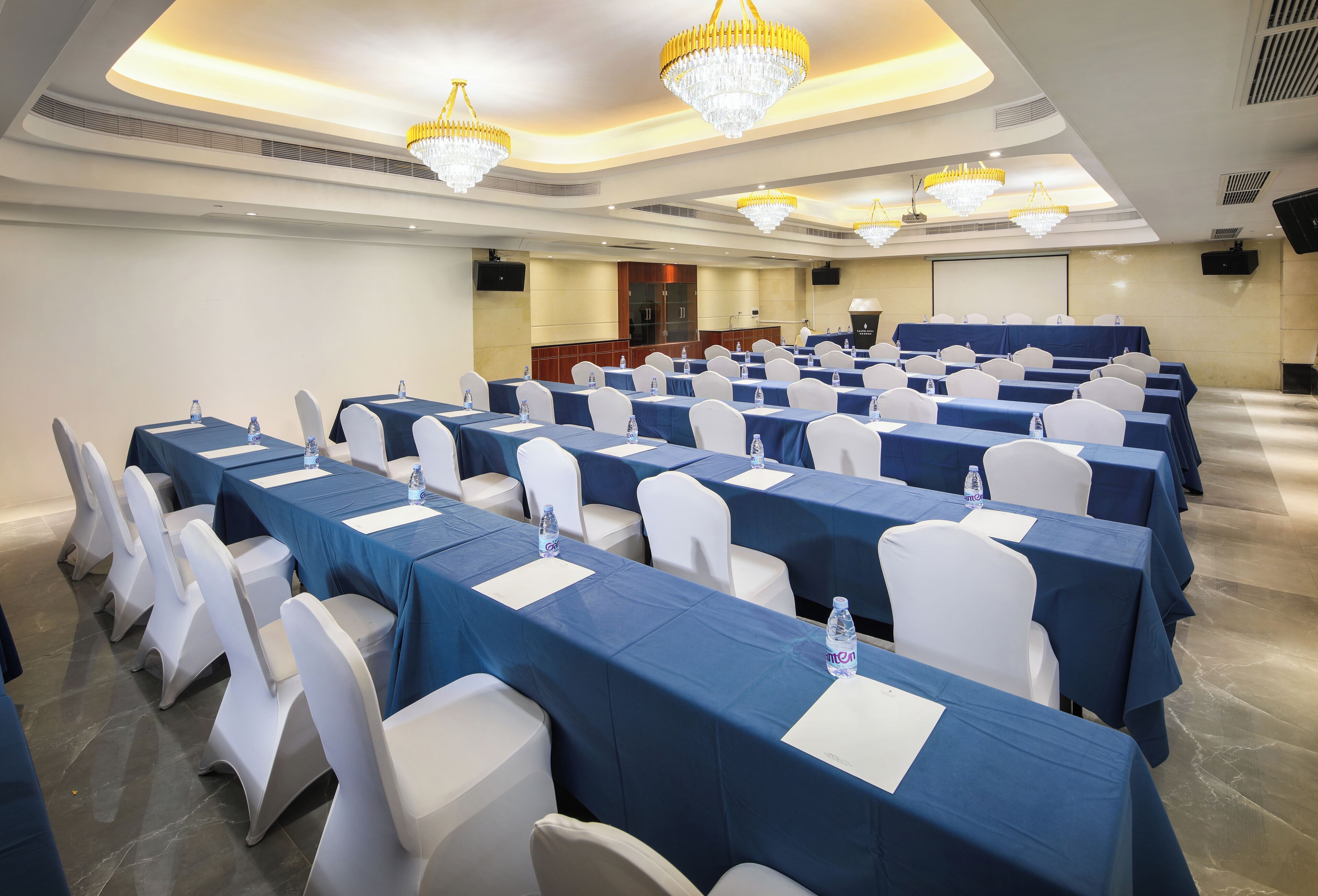 Un salón de conferencias con mesas dispuestas en filas, sillas blancas y manteles azules.