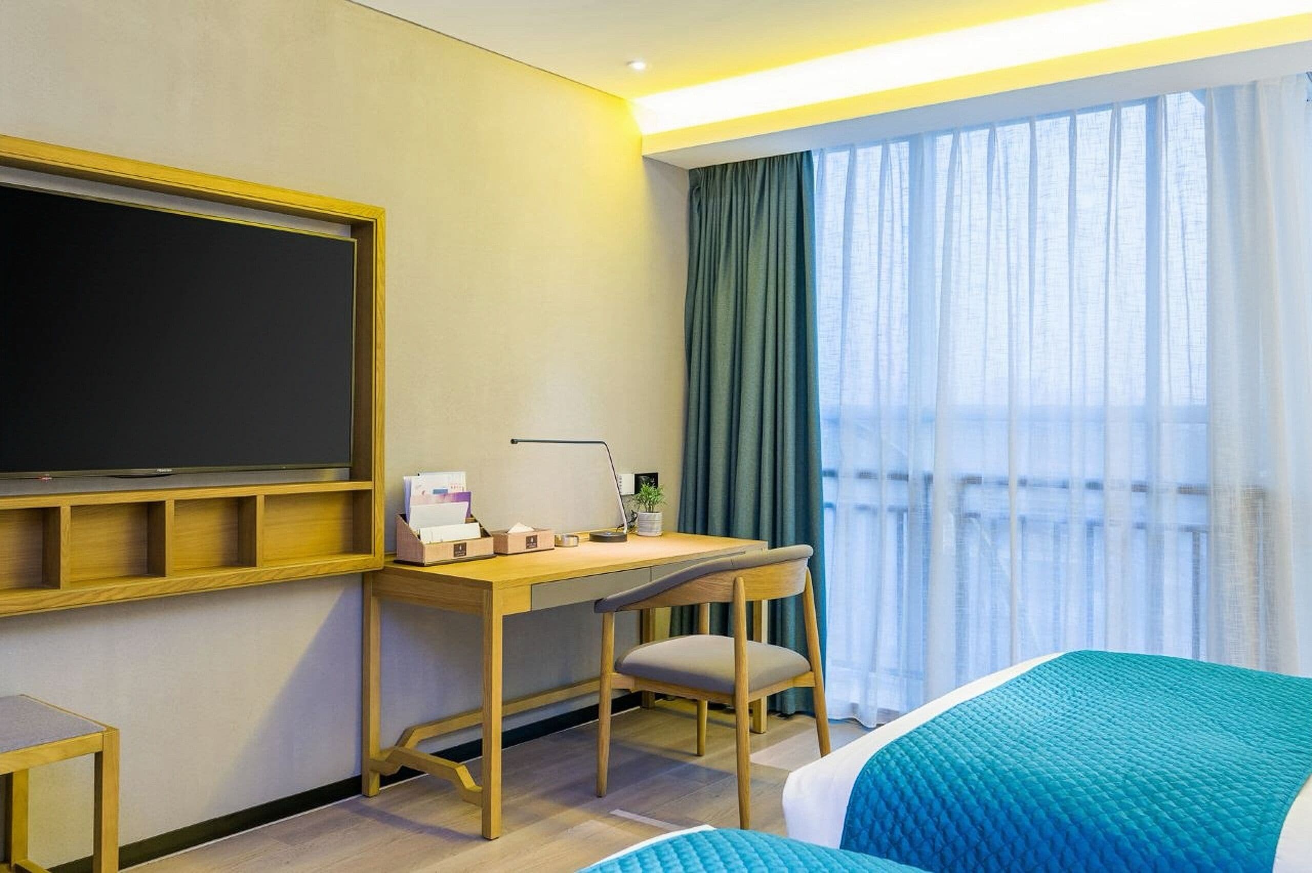 A hotel room with a bed, a desk with a chair, a television, and a large window with curtains.