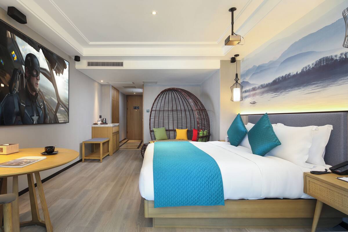 Quarto de hotel moderno com uma cama grande, uma mesa de jantar, uma pequena cozinha e um quadro grande na parede.