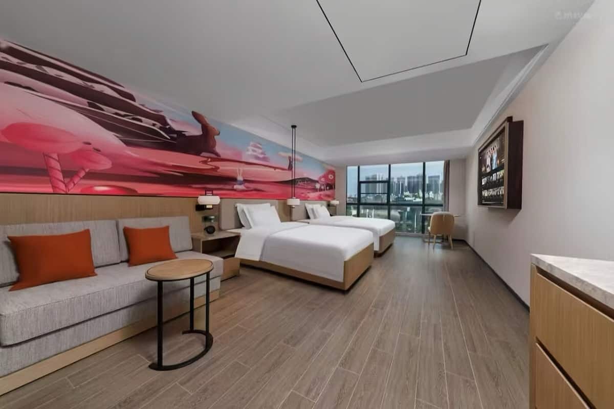 Quarto de hotel moderno com uma cama grande, um sofá, uma mesinha e vista para a cidade através de grandes janelas.