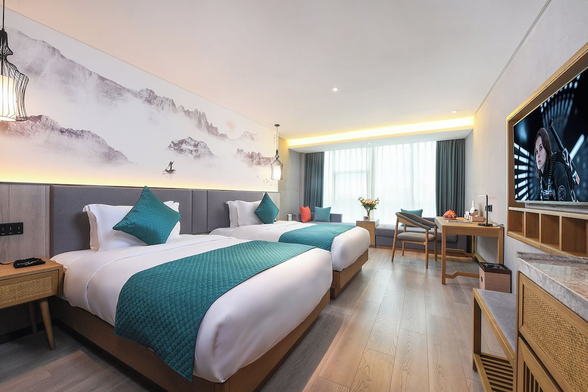 Quarto de hotel com duas camas, uma escrivaninha, televisão e uma janela grande com cortinas.