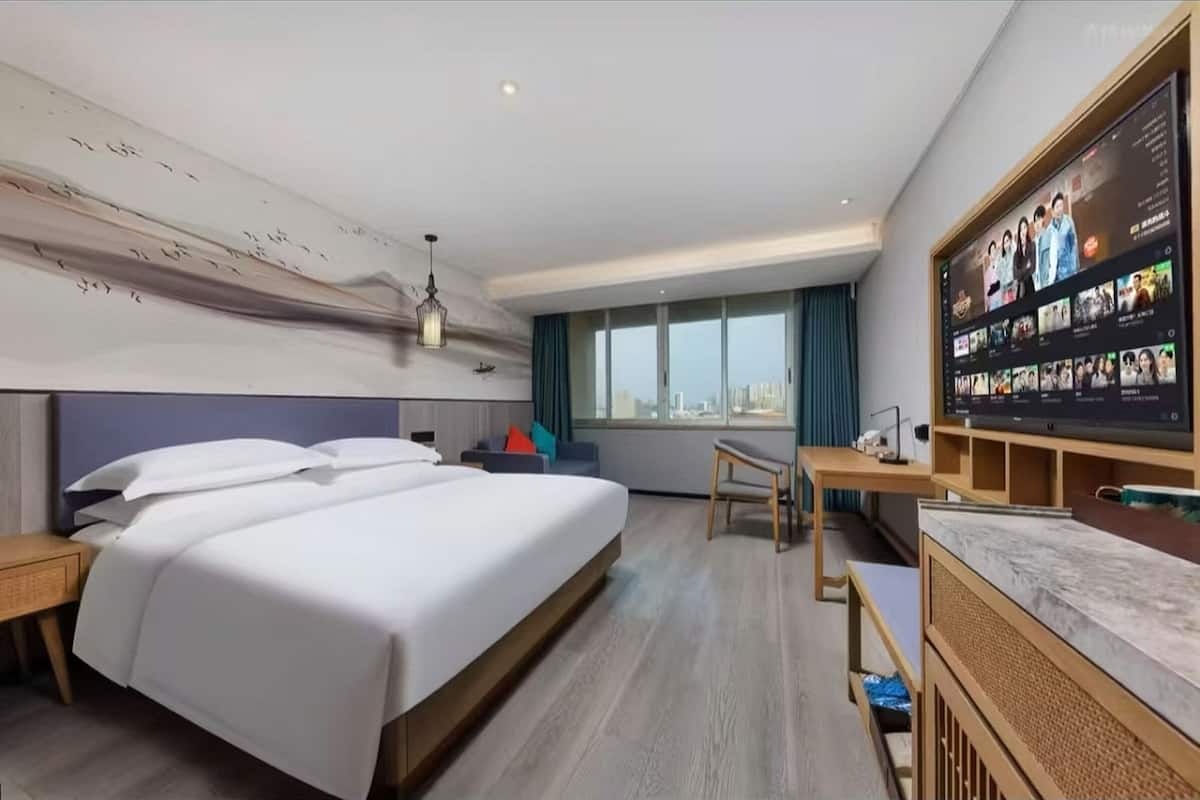 Quarto de hotel com uma cama grande, uma escrivaninha, televisão e janela com vista para a cidade.