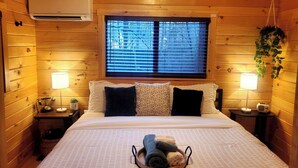 Un dormitorio tipo cabaña de madera con una cama, dos mesitas de noche, una ventana con persianas, aire acondicionado y una planta colgante.