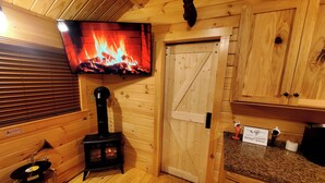 Interior de una cabaña de madera con chimenea, un televisor mostrando fuego, armarios de madera y un cartel que indica disponibilidad de WiFi.