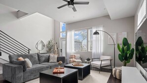 Een moderne woonkamer met een grijze bank, een zwarte salontafel en een witte fauteuil. Links een trap en rechts een groot raam met zonwering.