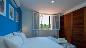 Une chambre avec un grand lit, une fenêtre avec des rideaux, une vue sur la verdure à l’extérieur et des volets en bois sur le côté droit.
