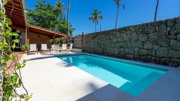Une piscine bordée d’un mur en pierre, avec des chaises longues et des palmiers en arrière-plan.