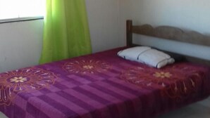Ein Einzelbett mit Holz-Kopfteil, eine violette Bettdecke mit Blumenmuster und ein grünlicher Vorhang, der teilweise zur Seite gezogen ist.