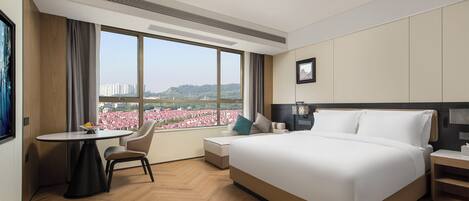 A hotel room with a large bed, a desk, and a chair. There is a window with a view of buildings and a cityscape.