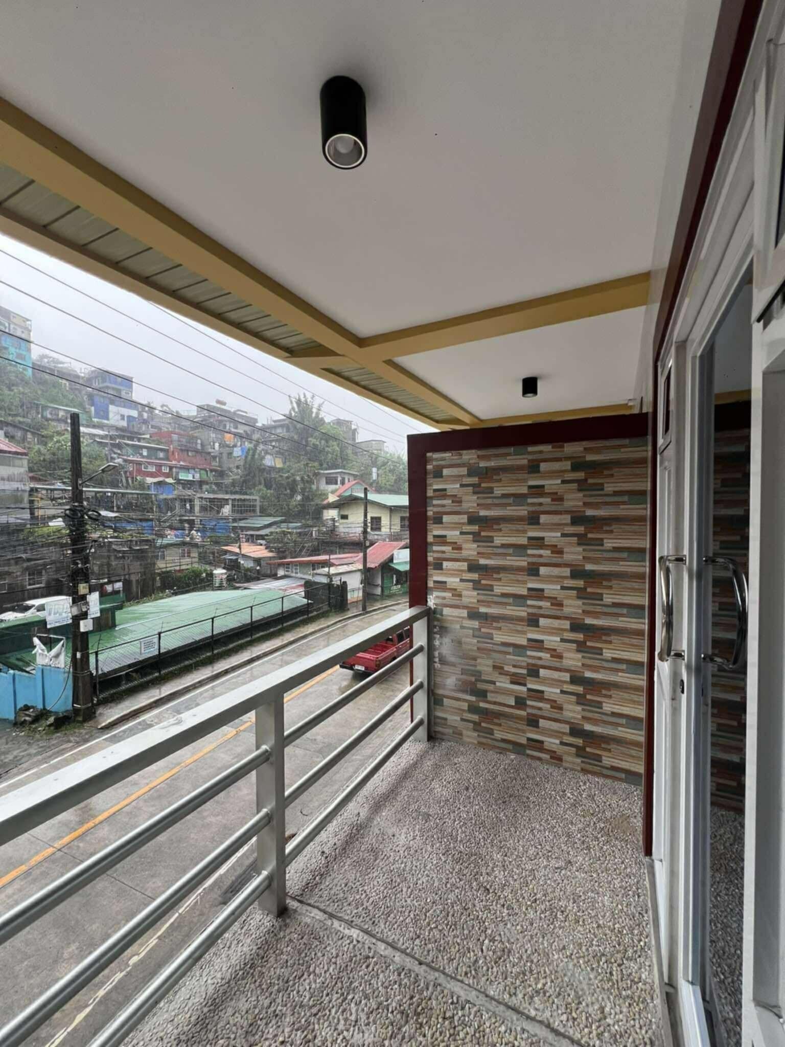 Un balcón con barandilla metálica, un muro con diseño y vista a un paisaje urbano con edificios y una carretera.