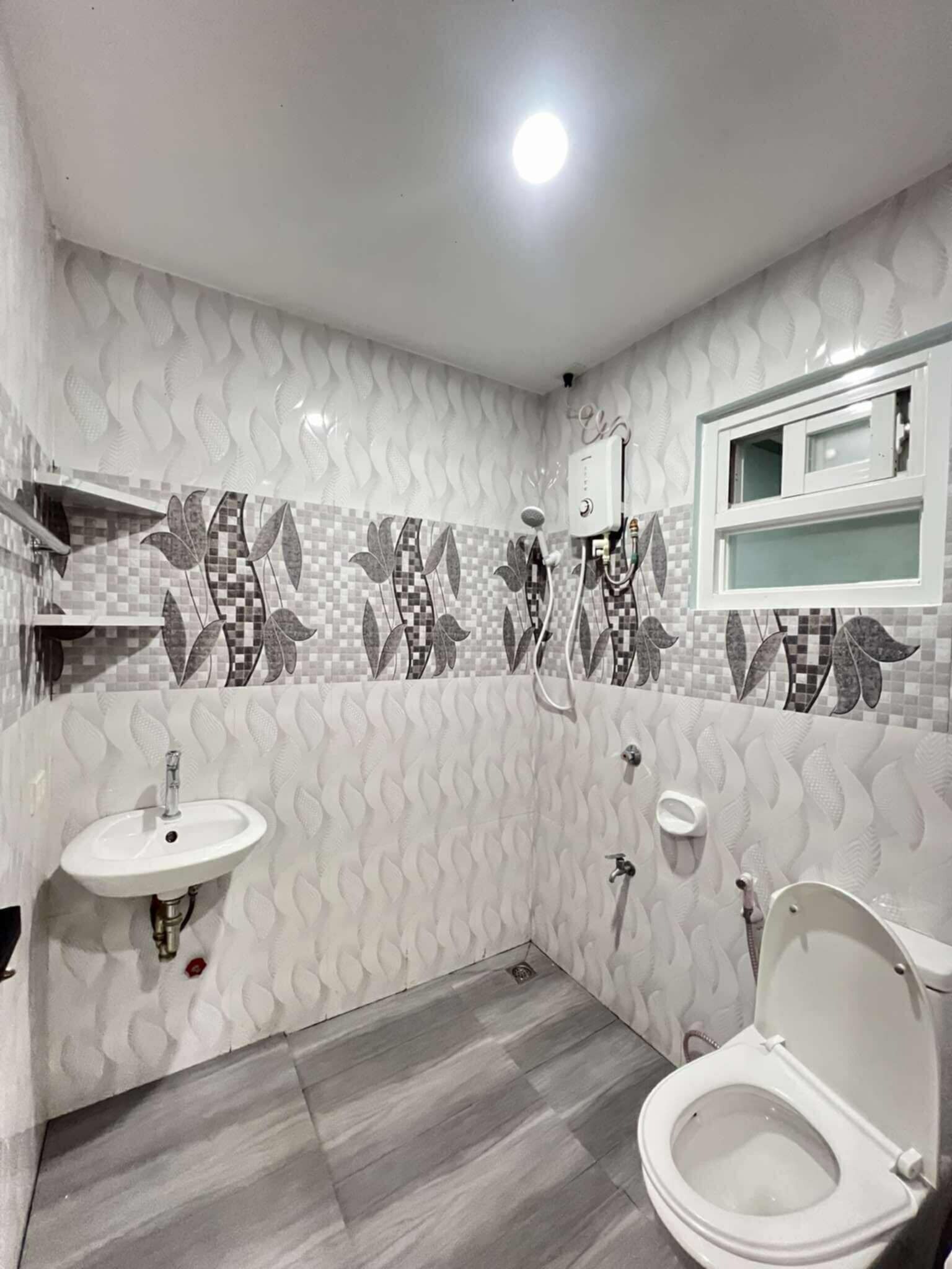 A bathroom with a white sink, toilet, and shower area, featuring patterned tiles and a window above the mirror.