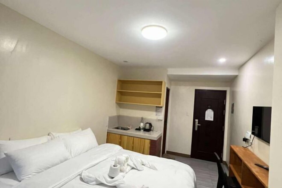 Ein Hotelzimmer mit Bett, einer kleinen Küchenzeile, einer Tür und einem Fernseher auf einem Holzständer.