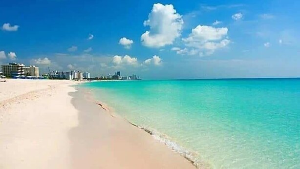 Una playa con agua turquesa cristalina y una orilla arenosa. Se observan edificios al fondo.