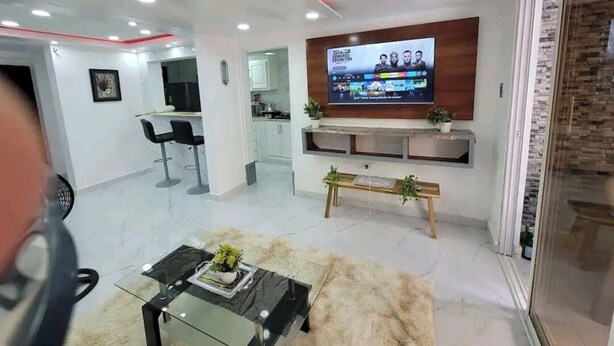 A modern living room with a flat-screen TV mounted on the wall, a glass coffee table, a fur rug, and a kitchen area in the background. - Obynb premium  (Santo Domingo)