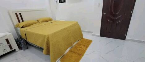 A bedroom with a bed, a white wall, an air conditioning unit, and a wooden door.