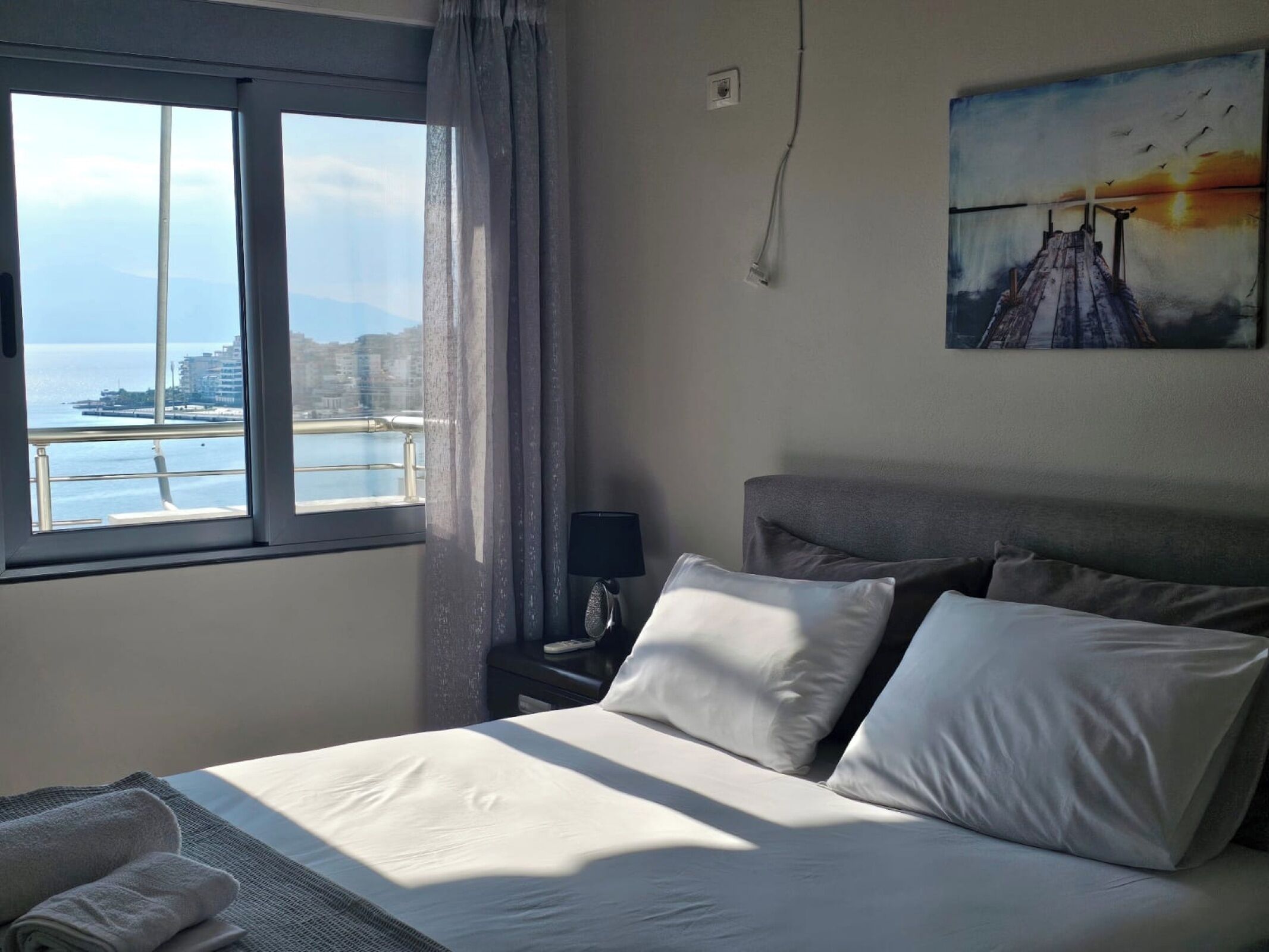Une chambre d’hôtel avec un lit, une table de chevet, une lampe, un tableau et une vue sur la mer et les bâtiments.