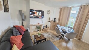 A modern living room with a black sofa, a TV mounted on the wall, a dining area with a table and chairs, and a small side table with a plant.
