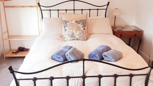 A bed with a metal frame, a wooden nightstand, a lamp, and a shelf with a bag and a book. - Stylish 2-Bed House in Stamford Town Centre (Stamford)