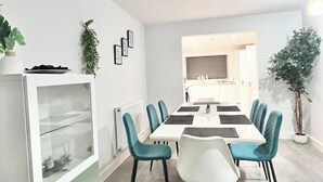 Et moderne spiseområde med et hvidt bord og turkise stole, et hvidt skab med glasdøre og en potteplante til venstre.