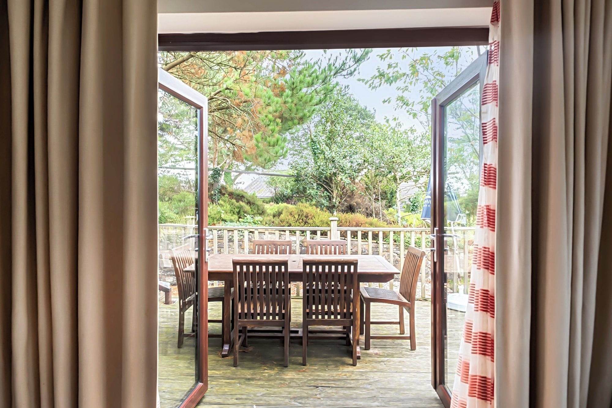 Una puerta corrediza de vidrio abierta que da a un patio exterior con una mesa y sillas de madera, rodeado de una terraza de madera y vistas a árboles y arbustos.