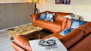 Een bruine leren bank met gekleurde kussens, een glazen salontafel en een wandlamp.