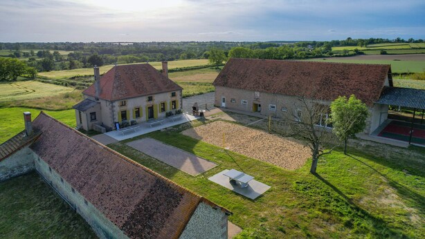 Un vaste domaine comprenant plusieurs bâtiments, dont une maison principale avec un toit rouge et une construction plus petite dotée d’un toit similaire. On y trouve une cour pavée, un banc et un arbre.