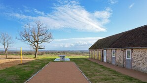 Un chemin mène à un bâtiment en pierre, surmonté d’un toit en tuiles, et entouré de pelouse et d’arbres.