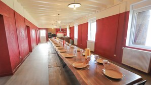 Une longue table à manger entourée de nombreuses chaises, des murs rouges et un coin cuisine à l’arrière-plan.