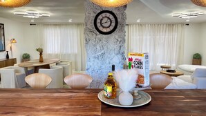 Un espace repas moderne avec une table en bois, des chaises blanches, une petite table ronde ornée d’un vase, une bouteille de spiritueux et une boîte de céréales Kellogg’s Corn Flakes.