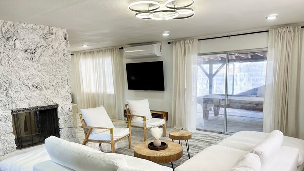 Un salon moderne avec un canapé blanc, deux fauteuils blancs, une cheminée, un téléviseur à écran plat et une grande fenêtre avec des rideaux.