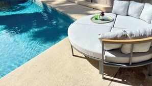 Un espace aménagé au bord de la piscine, avec un coin salon circulaire doté de coussins blancs et une petite table sur laquelle se trouvent une bouteille et un verre.