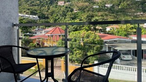 A balcony with two wicker chairs and a small round table, overlooking a hillside with houses and greenery.