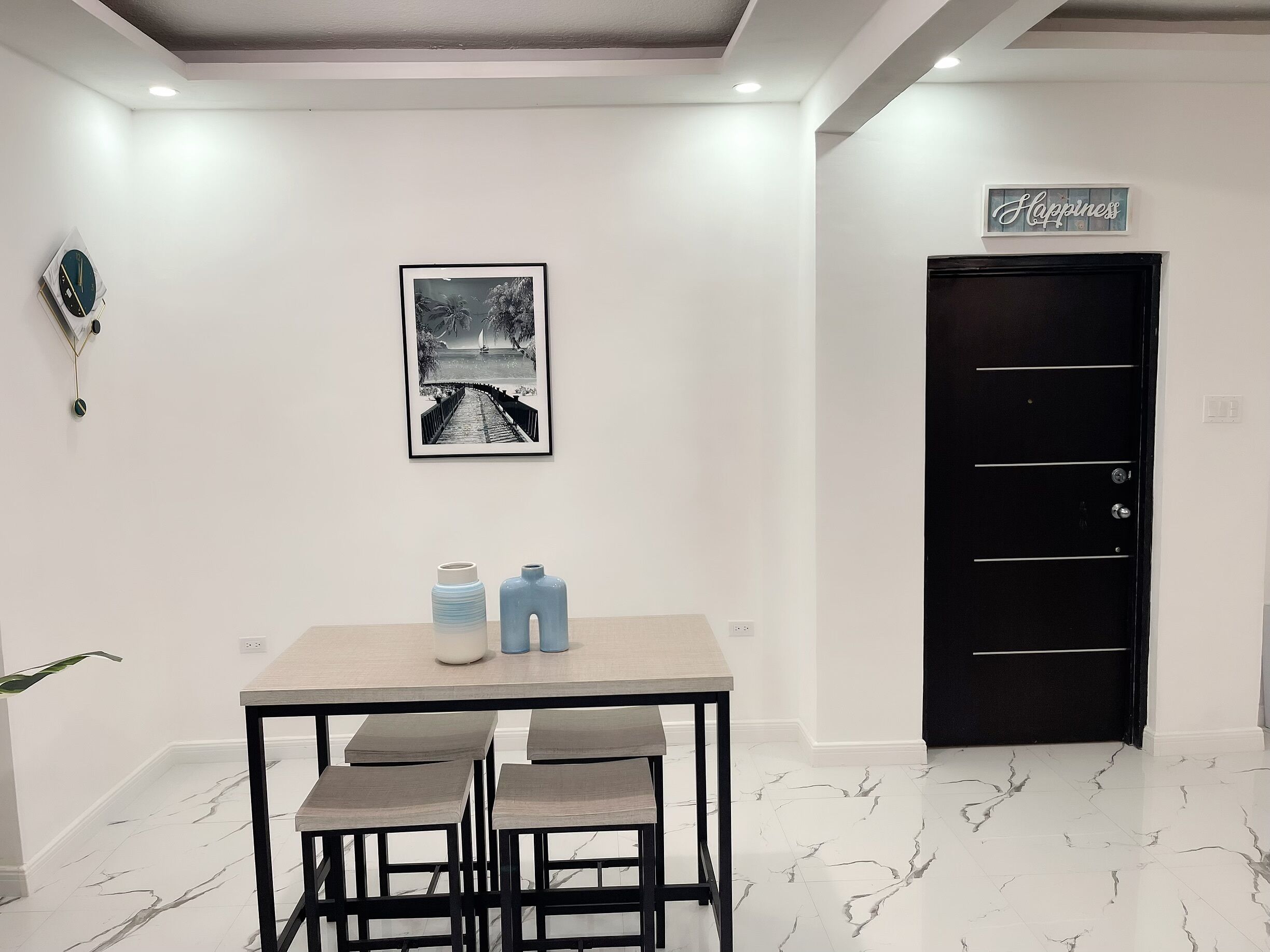 A dining area with a table and chairs, a framed picture on the wall, a black door, and a 'Happiness' sign.